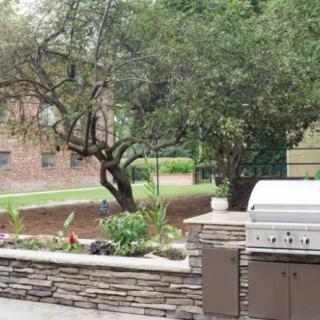 A patio area features a barbecue grill with a stone surround, set against a backdrop of trees and a brick wall.