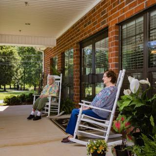 Two individuals enjoy a peaceful afternoon on a porch, seated in rocking chairs surrounded by greenery.
