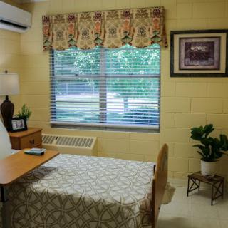 A cozy bedroom with a neatly made bed, a wooden table, and a window adorned with curtains, allowing natural light to illuminate the space.