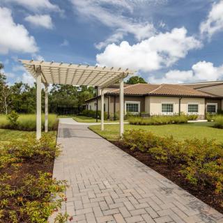 A scenic pathway with a pergola leads to a modern building surrounded by lush greenery under a partly cloudy sky.