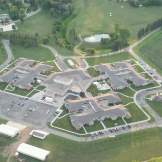 Aerial view of a building complex surrounded by green landscape and parking facilities.