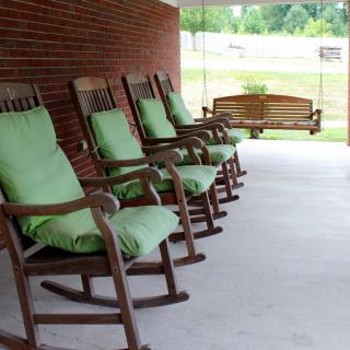A row of wooden rocking chairs with green cushions line a porch, accompanied by a swing in the background.