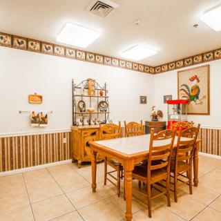A cozy kitchen dining area with a rustic rooster-themed decor, featuring wooden table and chairs, bright lighting, and decorative artwork.