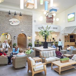 A spacious and well-lit lobby featuring comfortable seating areas, with several people gathered in conversation and a large decorative plant as the central focus.
