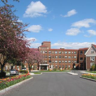 A view of a large hotel building with a circular driveway, surrounded by trees under a blue sky with clouds.