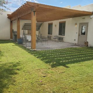 A cozy backyard featuring a shaded patio under a wooden pergola, surrounded by lush green grass.