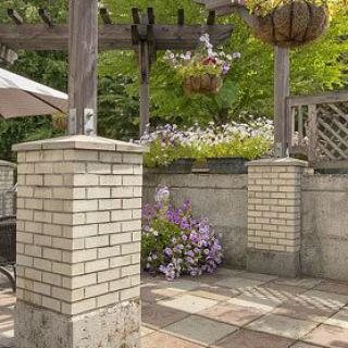 A serene patio area with brick pillars, hanging plants, and blooming flowers under a wooden pergola.