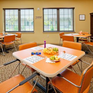 A cozy dining room setup with tables and chairs, featuring bingo cards and a fruit bowl under natural daylight.