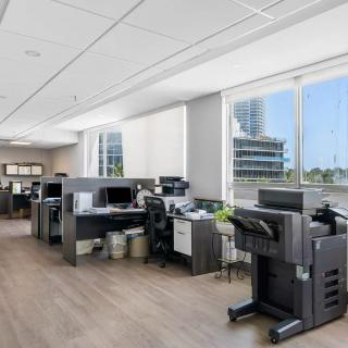 A modern office space with multiple desks, computers, and printers, featuring large windows allowing natural light to fill the room.