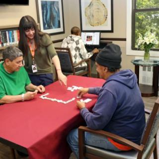 A group of people playing dominoes at a community center, with an assistant guiding the activity.