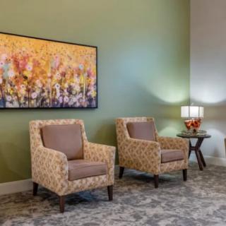 A cozy living room setup featuring two patterned armchairs, a side table with a lamp and flowers, and a vibrant painting on the wall.