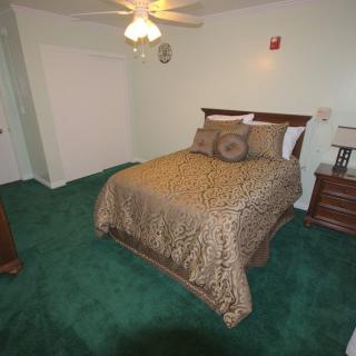 A cozy bedroom with a double bed, green carpet, wooden furniture, and a ceiling fan.