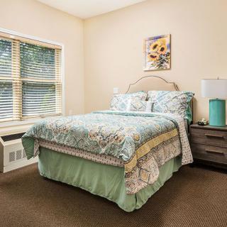 A cozy bedroom featuring a neatly made bed with patterned bedding, a window with blinds, and a nightstand in the corner.