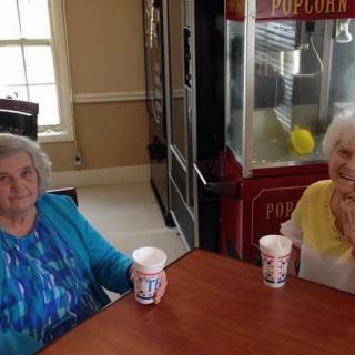 Two elderly women sitting at a table indoors with drinks, near a popcorn machine.