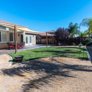 A sunny backyard with a mix of grass and sandy areas, bordered by trees and featuring a patio.