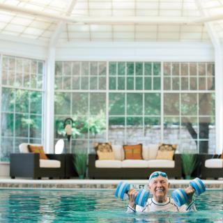 A senior woman doing water aerobics in an indoor pool with a relaxation area in the background.