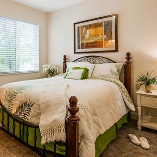 A cozy bedroom with a well-made bed, artwork on the wall, and a nightstand adorned with a plant and lamp.