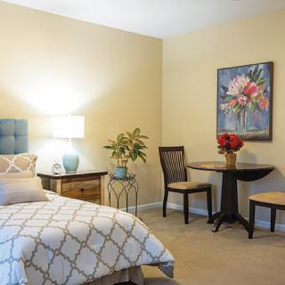 A cozy bedroom with a bed, bedside table, lamp, small round dining table with two chairs, decorative plants, and a floral artwork on the wall.