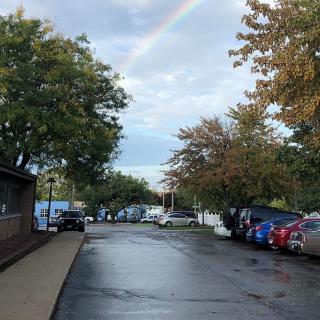 A rainbow stretches over a parking lot with wet pavement and cars, surrounded by trees on an overcast day.