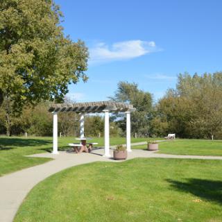 A serene park setting featuring a white pavilion surrounded by lush green grass and trees under a clear blue sky.