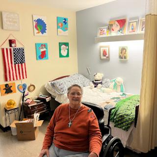 A person seated in a wheelchair in a warmly decorated room featuring artwork and personal touches.