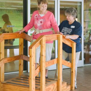 A caregiver assists an elderly individual as they practice walking on a wooden therapy ramp.