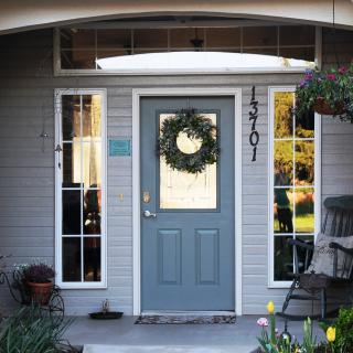 A cozy front porch with a gray door adorned with a wreath, flanked by windows and surrounded by plants.