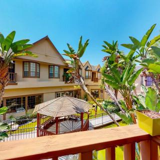 A picturesque view from a balcony overlooking a charming residential area with tropical plants and an outdoor gazebo under a clear, sunny sky.