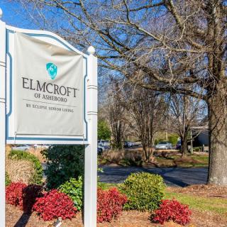 A sign for Elmcroft of Roswell is situated in a landscaped area with trees and shrubs, next to a parking lot.