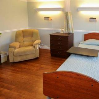 A well-lit room with two beds, a comfortable chair, and wooden flooring, typical of a nursing home or hospital setting.