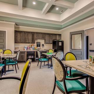 A well-lit dining area with colorful chairs and tables, featuring a kitchenette in the background.