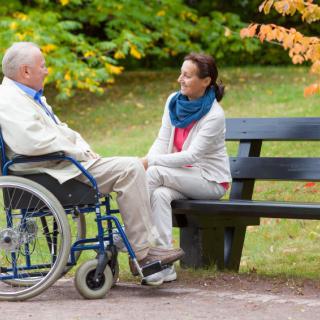 An elderly man in a wheelchair and a woman having a conversation on a park bench during autumn.