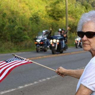 An elderly woman waves an American flag by the roadside as motorcycles pass by.
