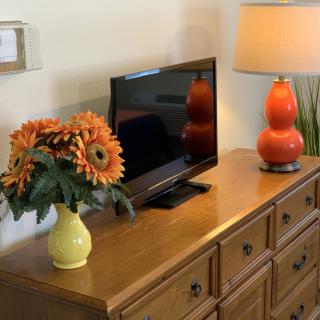 A cozy living room setup with a wooden dresser, television, orange lamp, and a vase of flowers.