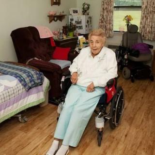 An elderly person sitting in a wheelchair in a cozy room with a bed and armchair.