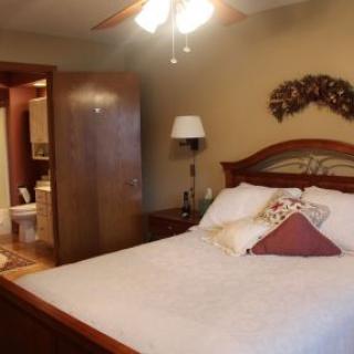 A cozy bedroom with wooden furniture, featuring a neatly made bed adorned with decorative pillows and a ceiling fan overhead.