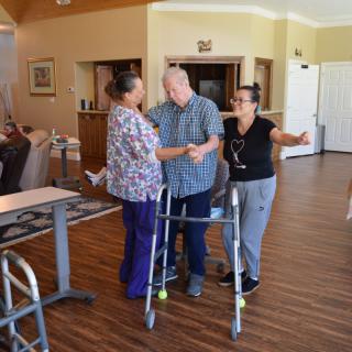 Caregivers assist an elderly man using a walker in a nursing home setting.