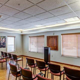 A small conference room with arranged chairs and a podium, featuring large windows and overhead lighting.