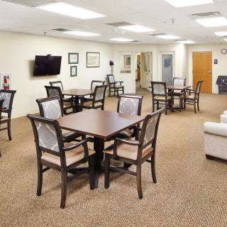 A well-lit dining area with multiple tables and chairs arranged on a carpeted floor, featuring a wall-mounted television.