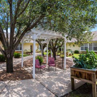 A peaceful garden setting featuring a white gazebo with two pink chairs underneath, surrounded by lush trees and greenery.