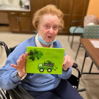 An elderly woman in a wheelchair proudly displays her colorful painting indoors.