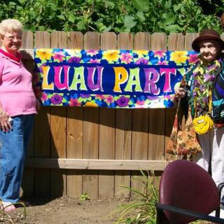 Two people standing next to a colorful 'Luau Party' banner outdoors.