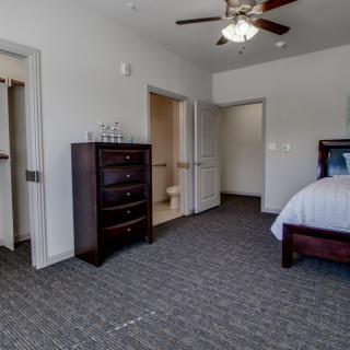 A spacious bedroom with a bed, dresser, and ceiling fan, featuring carpeted flooring and open doorways.