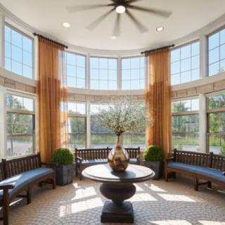 A bright living space with large windows, elegant curtains, and a central round table adorned with a decorative vase. The seating area offers a cozy and inviting atmosphere.