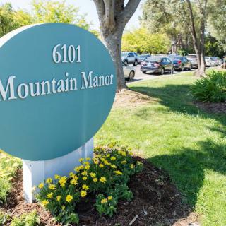 A sign reading '6101 Mountain Manor' is displayed in a landscaped area with trees and parked cars nearby.