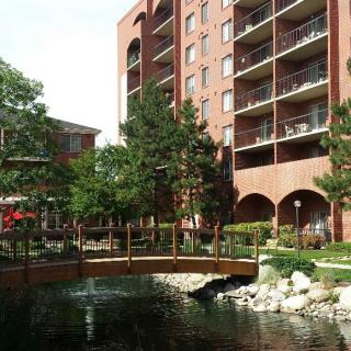 A scenic view of an apartment building with a small bridge over a pond surrounded by trees and greenery.