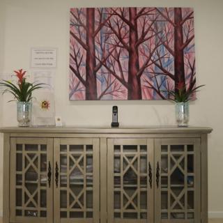 A decorated cabinet sits below a colorful painting of trees, flanked by plants.