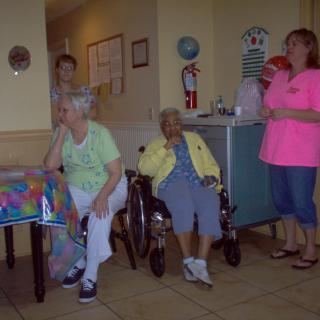 A group of elderly individuals celebrate a birthday indoors, with balloons and gifts.