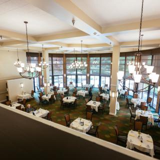 A spacious restaurant dining room with elegantly set tables and large windows, viewed from above.