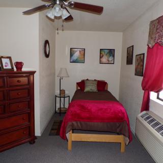 A cozy bedroom with a single bed featuring red bedding, a wooden dresser, a lamp, and artwork on the walls.